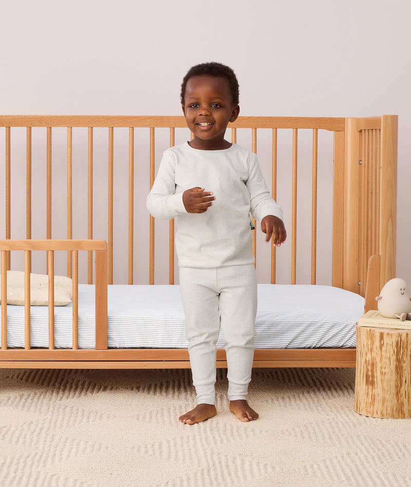 A toddler wearing ergoPouch Long Sleeve Pyjamas standing next to a toddler bed.