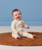 A happy baby sitting on a brown quilted mat, wearing a patterned cream Long Sleeve Layer.