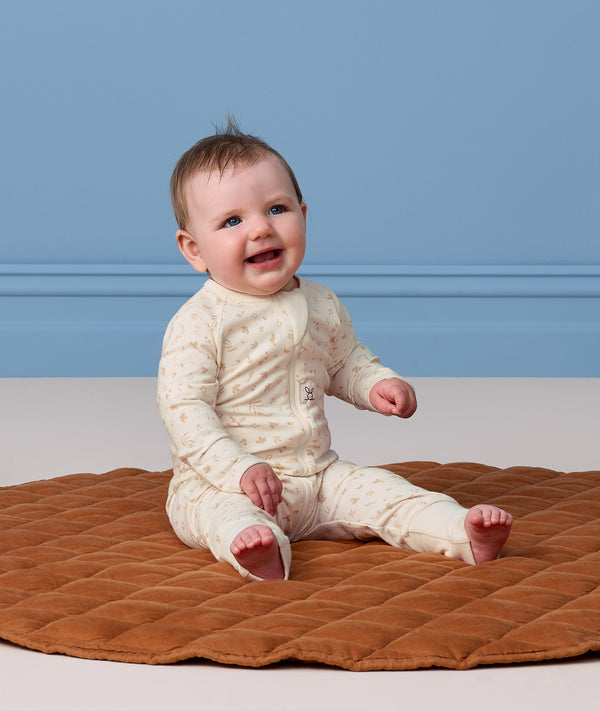 A happy baby sitting on a brown quilted mat, wearing a patterned cream Long Sleeve Layer.