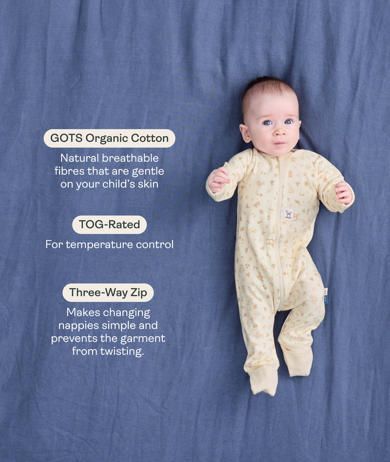 Baby lying on a blue blanket wearing a cream patterned Long Sleeve Layer with text highlighting GOTS Organic Cotton, TOG Ratings, and Three-way zipper.