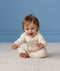 A happy baby sitting on a cream textured rug, wearing a patterned cream Long Sleeve Layer with hands outstretched toward the camera.