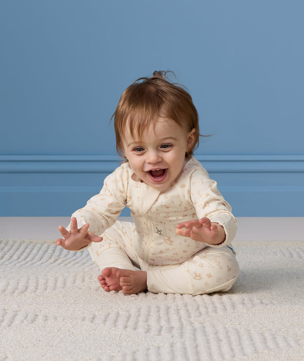 A happy baby sitting on a cream textured rug, wearing a patterned cream Long Sleeve Layer with hands outstretched toward the camera.