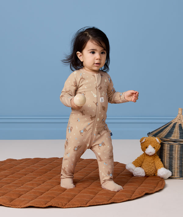 A toddle stands on a round, brown quilted rug in front of a solid blue wall. The child is wearing a beige printed Long Sleeve Layer and holding a smooth wooden ball, with a plush stuffed animal and a woven storage basket on the floor nearby.