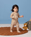 A toddle stands on a round, brown quilted rug in front of a solid blue wall. The child is wearing a beige printed Long Sleeve Layer and holding a smooth wooden ball, with a plush stuffed animal and a woven storage basket on the floor nearby.