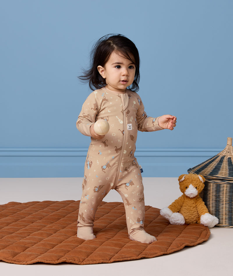A toddle stands on a round, brown quilted rug in front of a solid blue wall. The child is wearing a beige printed Long Sleeve Layer and holding a smooth wooden ball, with a plush stuffed animal and a woven storage basket on the floor nearby.