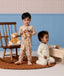 Two toddlers in patterned Long Sleeve Layers play on a brown rug in a nursery. One stands holding wooden binoculars next to a rocking chair, while the other kneels by a wooden toy chest.