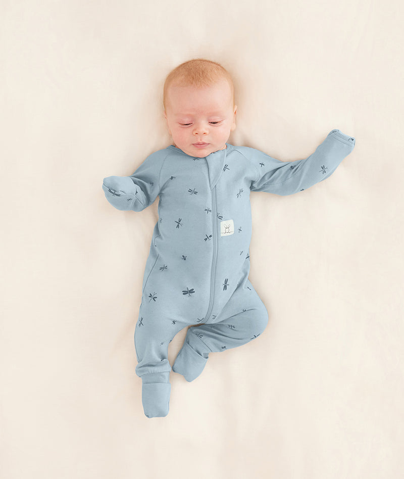 Baby is lying on a neutral coloured blanket, wearing a blue Long Sleeve Layer with its hand and foot cuffs folded over.