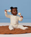 Baby sitting on a quilted brown mat against a light blue background, wearing a cream patterned Long Sleeve Layer, holding a wooden maraca in each hand.