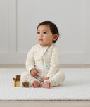 Infant sits on a rug wearing an ergoPouch Long Sleeve Layer in Oatmeal Marle.