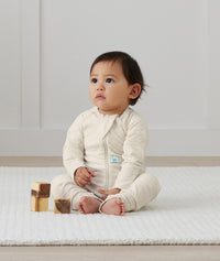 Infant sits on a rug wearing an ergoPouch Long Sleeve Layer in Oatmeal Marle.