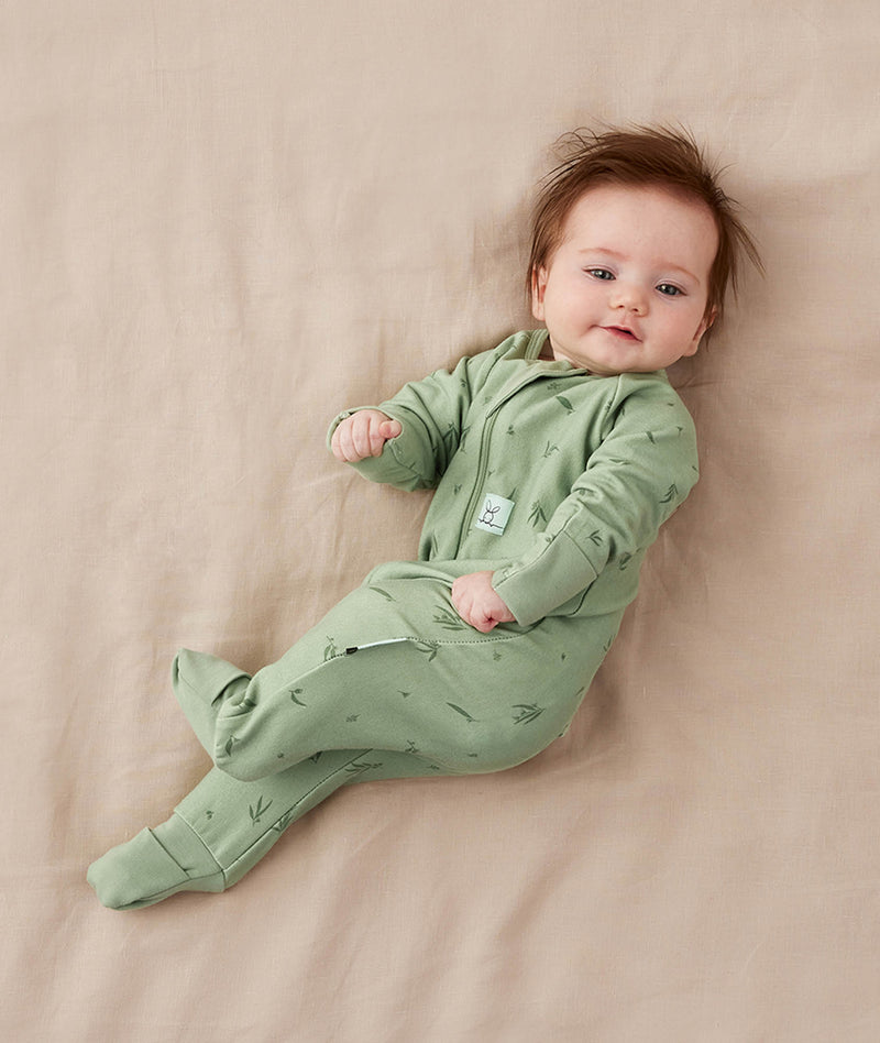 Baby lying on a soft pink blanket, wearing a green ergoPouch Long Sleeve Layer with its foot cuffs folded over for extra warmth and protection.
