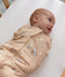 Smiling baby wrapped in a tan patterned Cocoon Swaddle Bag, lying on a blue-and-white pinstriped bassinet sheet.