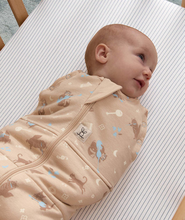 Smiling baby wrapped in a tan patterned Cocoon Swaddle Bag, lying on a blue-and-white pinstriped bassinet sheet.