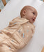 Smiling baby wrapped in a tan patterned Cocoon Swaddle Bag, lying on a blue-and-white pinstriped bassinet sheet.