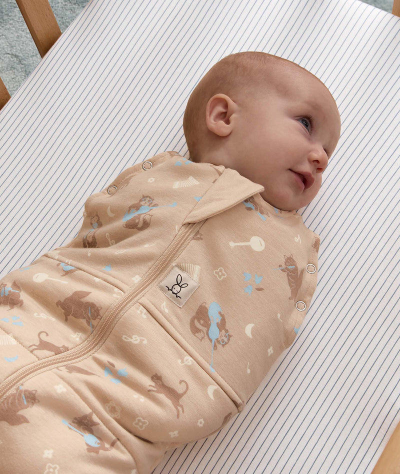 Smiling baby wrapped in a tan patterned Cocoon Swaddle Bag, lying on a blue-and-white pinstriped bassinet sheet.