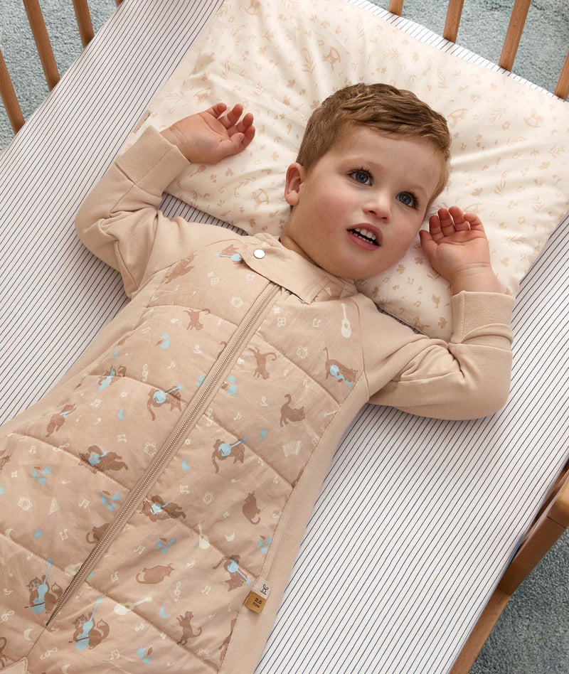 Toddler lying on his back in a wooden crib, wearing a beige sleep suit bag with his head on a matching toddler pillow.