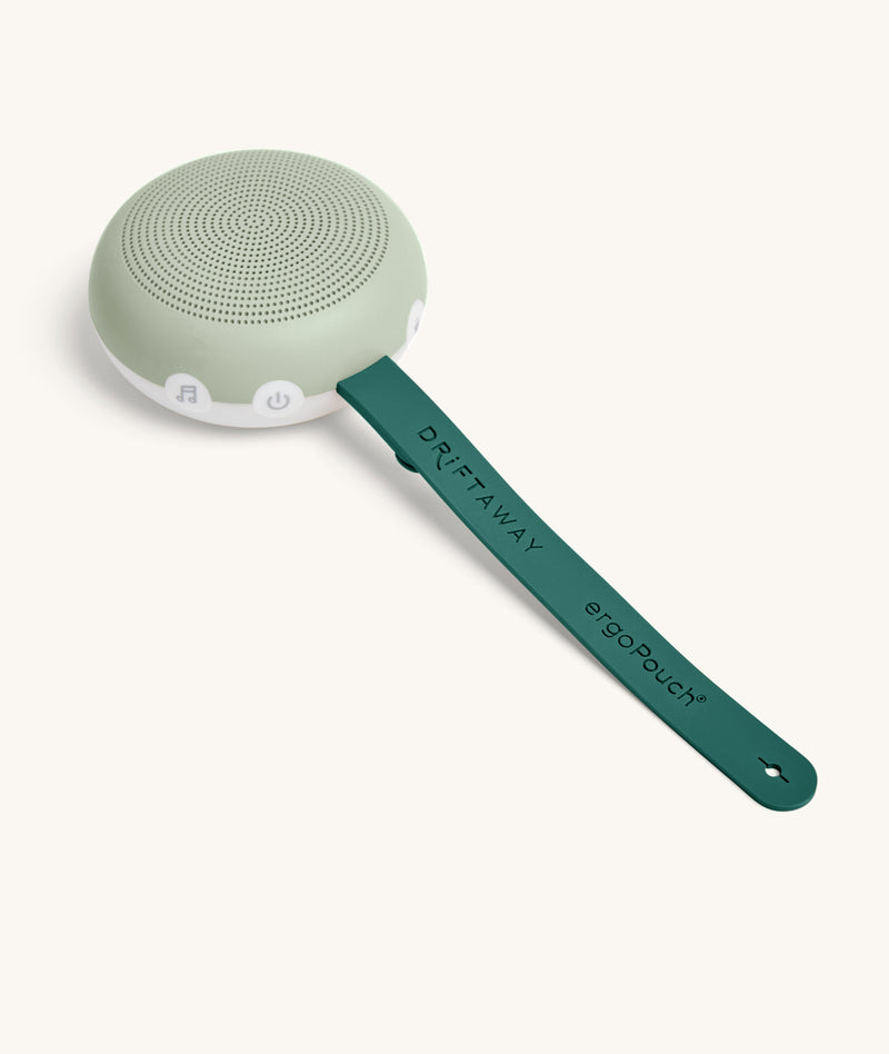 A sage green and white circular white noise machine with speaker holes on top and control buttons on the side sits attached to a dark green strap labeled “DRIFTAWAY ergoPouch®” on a white background.