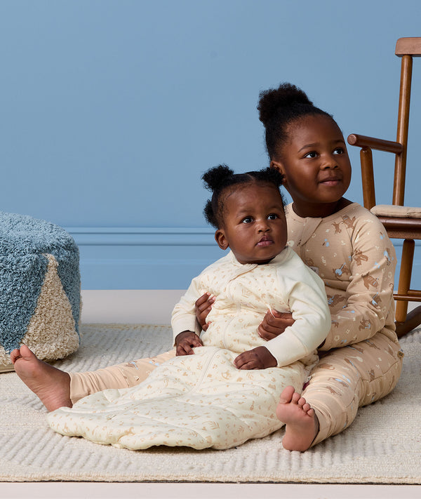 A preschooler sitting on a rug, wearing a Sleep Onesie, hugging a baby who is wearing a cream pattern-printed long-sleeve Sleeping Bag.
