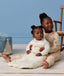 A preschooler sitting on a rug, wearing a Sleep Onesie, hugging a baby who is wearing a cream pattern-printed long-sleeve Sleeping Bag.
