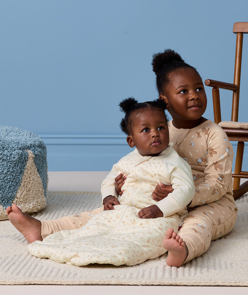 A preschooler sitting on a rug, wearing a Sleep Onesie, hugging a baby who is wearing a cream pattern-printed long-sleeve Sleeping Bag.