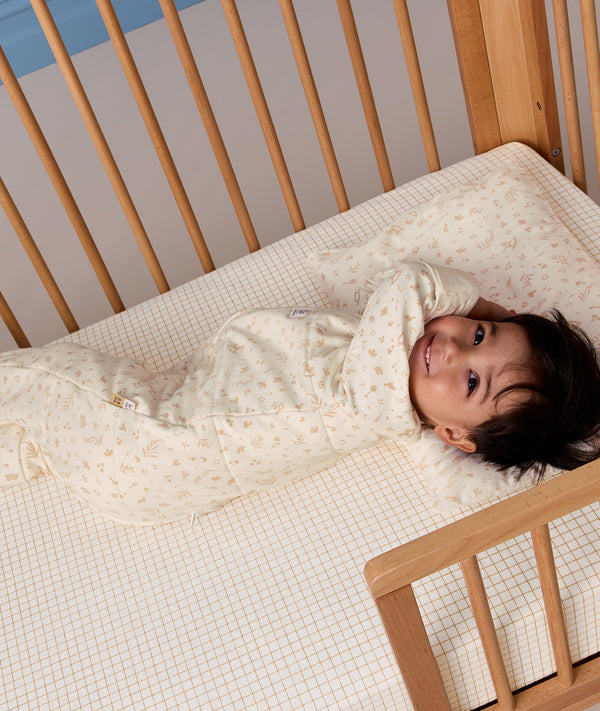 Toddler lying on their back in a wooden toddler bed, resting their head on a small matching pillow while wearing a cream patterned sleep onesie.