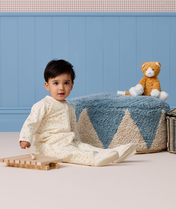 Toddler sitting on the floor in a cream patterned Sleep Onesie, playing with a wooden xylophone next to a blue pouf and plush cat.