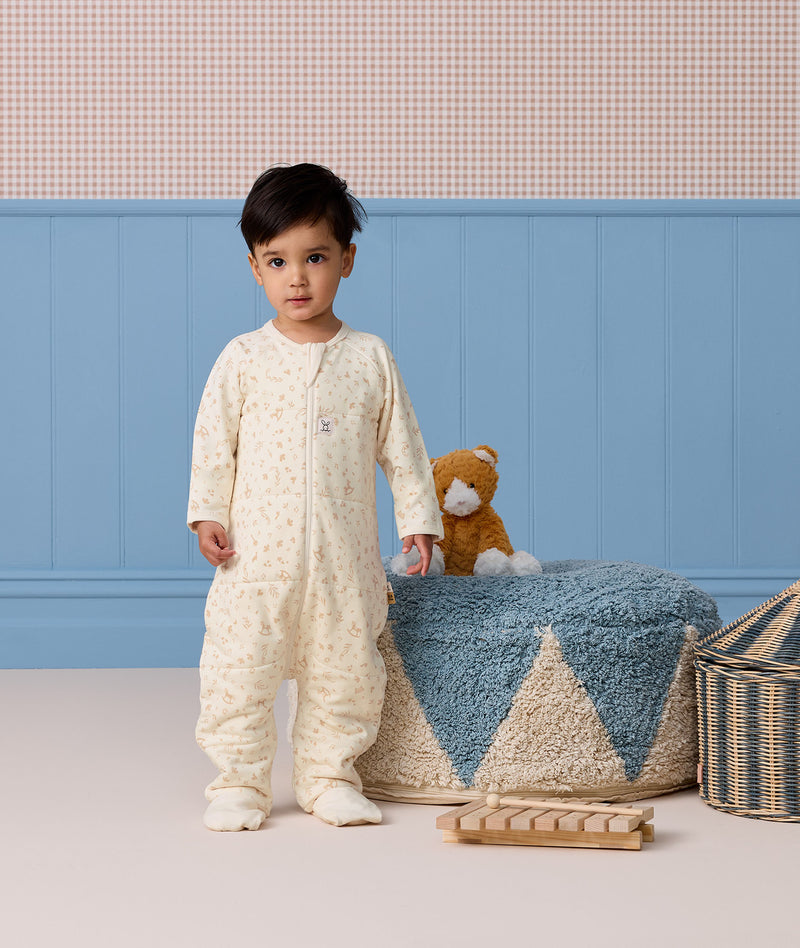 Toddler wearing a cream patterned Sleep Onesie with covered feet standing near a wooden toy xylophone and a plush cat.