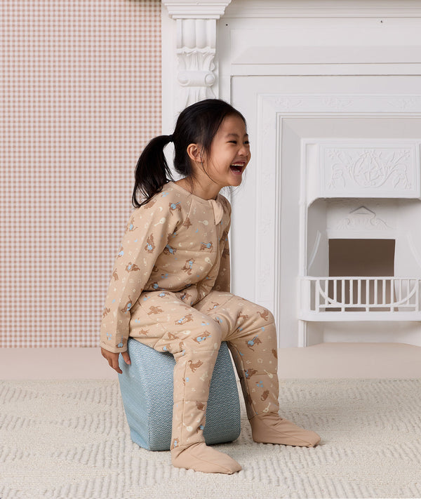 Laughing young girl sitting on a blue pouf, wearing a beige patterned Sleep Onesie with built-in foot cuffs.