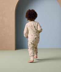 A toddler walks away from the camera wearing an ergoPouch Sleep Onesie with long sleeves and full-length legs. The onesie features an all-over animal print and a snug fit with foot cuffs, offering cozy comfort and flexibility for movement.