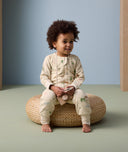 A toddler sits on a woven cushion wearing an ergoPouch Sleep Onesie decorated with colorful animal prints. The onesie features long sleeves, full-length legs, and a front zipper for easy wear.