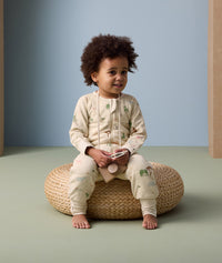 A toddler sits on a woven cushion wearing an ergoPouch Sleep Onesie decorated with colorful animal prints. The onesie features long sleeves, full-length legs, and a front zipper for easy wear.