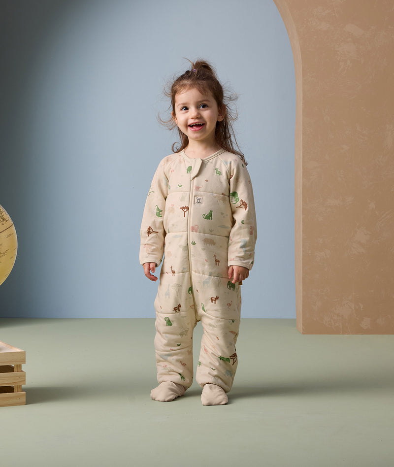 A smiling toddler stands in an ergoPouch Sleep Onesie decorated with playful animal prints. The onesie features long sleeves, a front zipper, and footed cuffs, designed to provide cozy warmth and unrestricted movement for sleep or play.