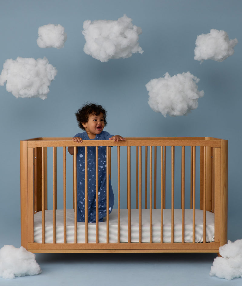 Child standing in a wooden crib wearing an ergoPouch Sleep Suit Bag 1.0 TOG Starbeam, a sleeping bag that converts to a sleep suit with legs.