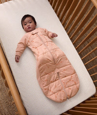 Baby lying in a wooden crib, wearing a peach-colored padded sleep suit bag with a front zipper and subtle animal print details.