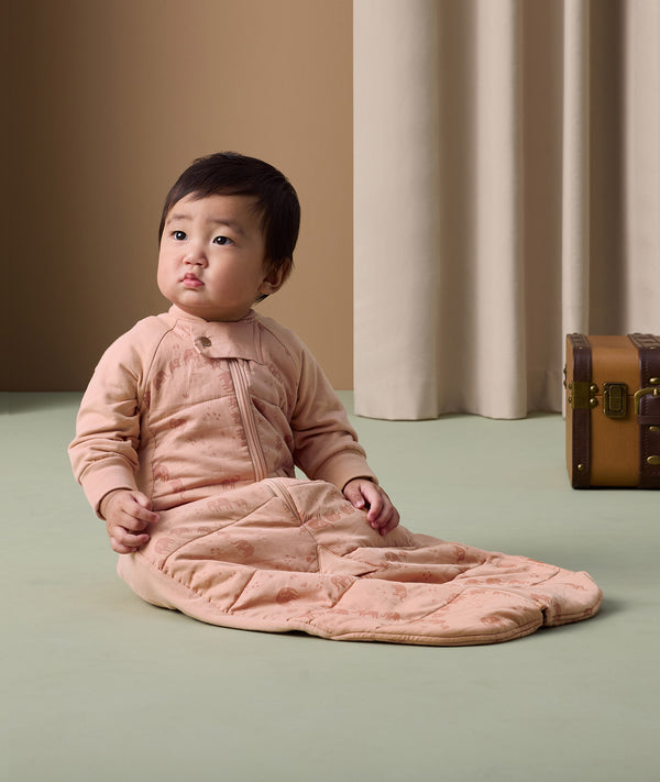 Baby sitting upright on a soft green floor, wearing a peach-colored padded sleep suit bag with a front zipper and subtle animal print.
