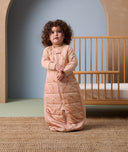 Toddler standing on a woven rug in front of a wooden crib, wearing a peach-colored sleep suit bag with a front zipper and subtle animal print details, in a softly lit nursery setting.