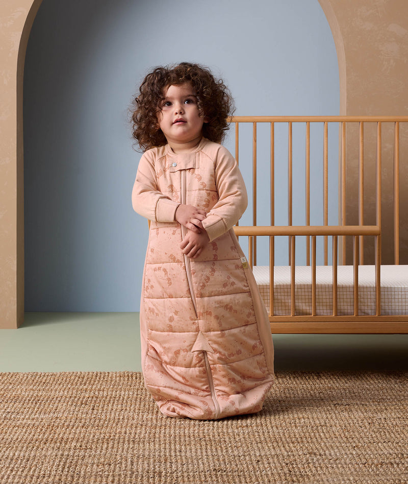 Toddler standing on a woven rug in front of a wooden crib, wearing a peach-colored sleep suit bag with a front zipper and subtle animal print details, in a softly lit nursery setting.