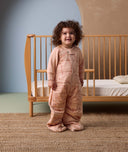 Smiling toddler standing in front of a wooden crib, wearing a peach-colored sleep suit with feet and a front zipper, decorated with a subtle animal pattern, in a cozy nursery setting.