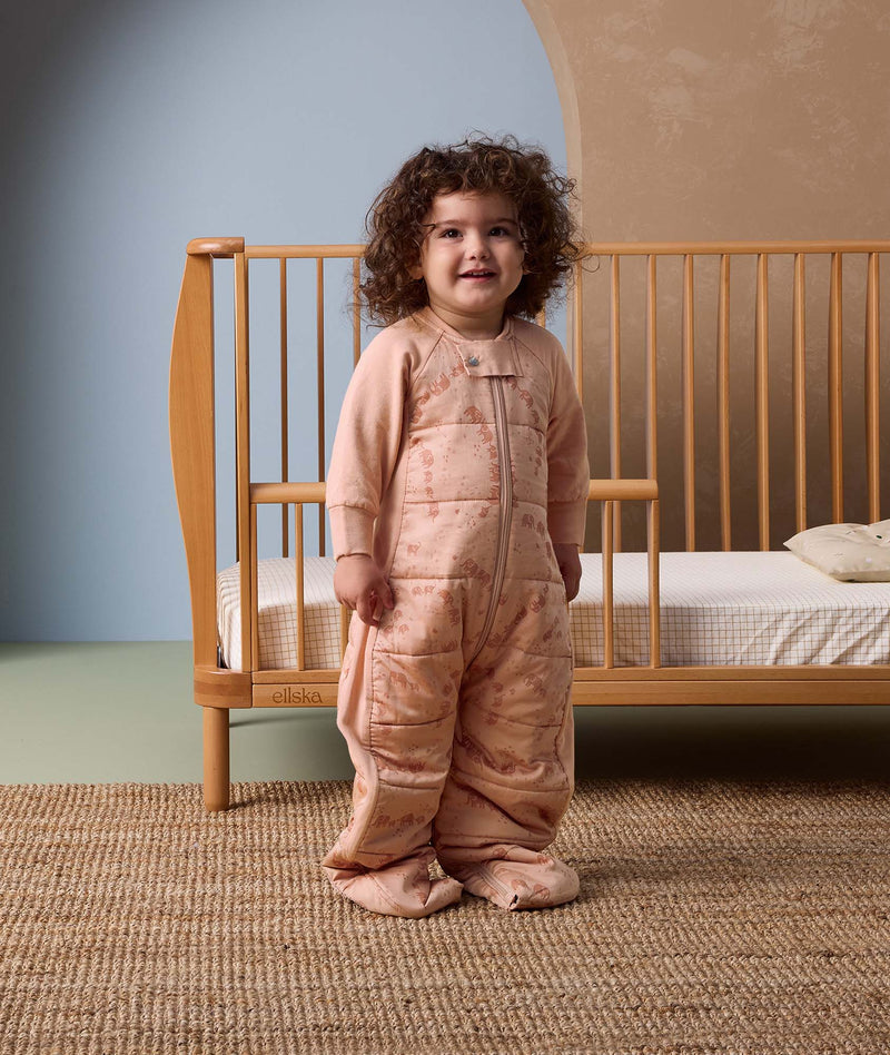Smiling toddler standing in front of a wooden crib, wearing a peach-colored sleep suit with feet and a front zipper, decorated with a subtle animal pattern, in a cozy nursery setting.
