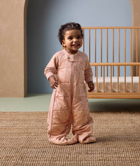 Smiling toddler standing in front of a wooden crib, wearing a peach-colored sleep suit with feet and a front zipper, decorated with a subtle animal pattern, in a cozy nursery setting.
