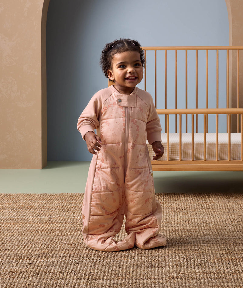 Smiling toddler standing in front of a wooden crib, wearing a peach-colored sleep suit with feet and a front zipper, decorated with a subtle animal pattern, in a cozy nursery setting.