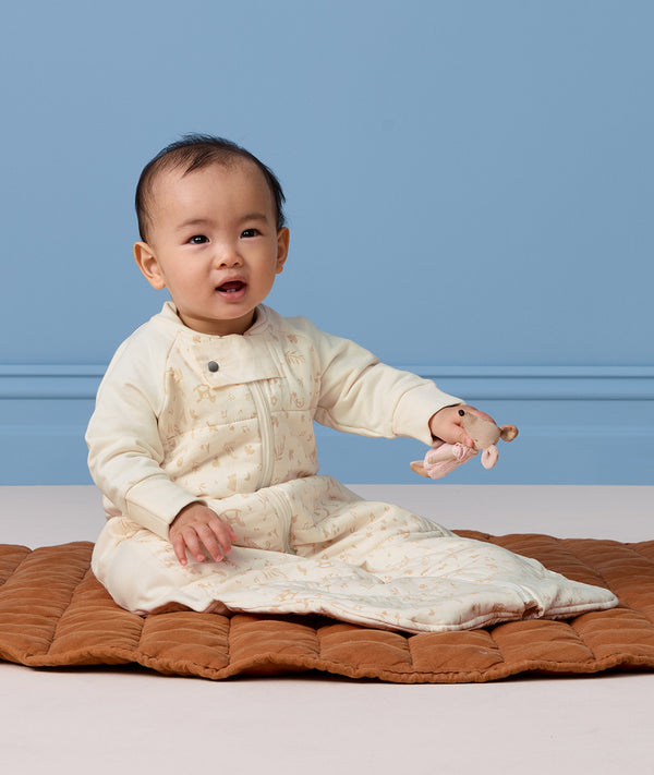 Baby sitting on a brown quilted mat wearing a cream patterned sleep suit bag while holding a small mouse toy.