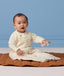 Baby sitting on a brown quilted mat wearing a cream patterned sleep suit bag while holding a small mouse toy.