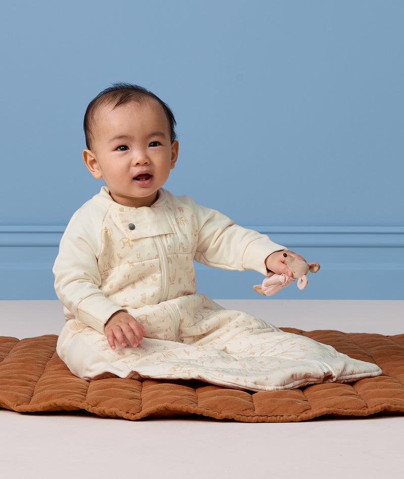 Baby sitting on a brown quilted mat wearing a cream patterned sleep suit bag while holding a small mouse toy.