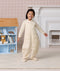 Smiling preschooler wearing a cream-colored patterned Sleep Suit Bag, standing with arms slightly outstretched next to a dollhouse.