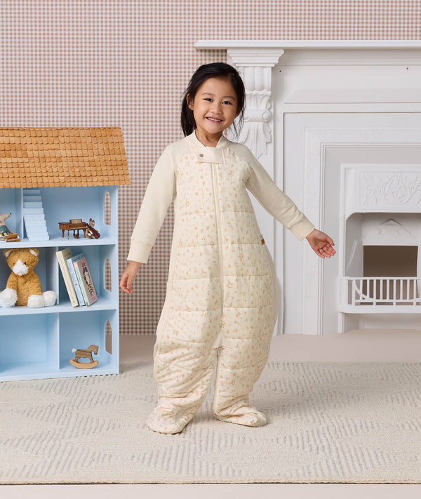 Smiling preschooler wearing a cream-colored patterned Sleep Suit Bag, standing with arms slightly outstretched next to a dollhouse.
