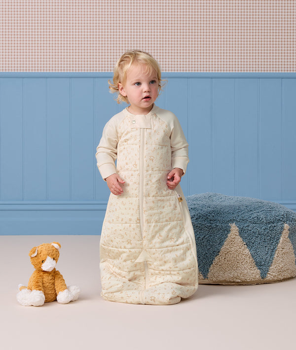 Toddler wearing a cream patterned sleep suit bag, standing in a room near a plush cat toy and a blue pouf.