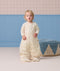 Toddler wearing a cream patterned sleep suit bag with legs, standing in a room near a blue pouf.