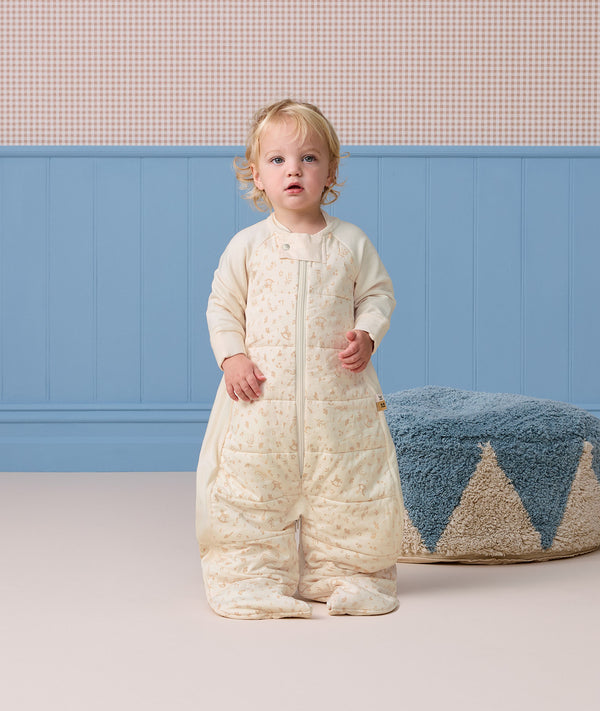 Toddler wearing a cream patterned sleep suit bag with legs, standing in a room near a blue pouf.