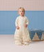 Toddler wearing a cream patterned sleep suit bag with legs, standing in a room near a blue pouf.